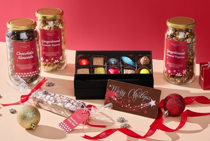 Jars of Christmas sweets and a box of chocolates with ribbons and baubles on a table against a red background.