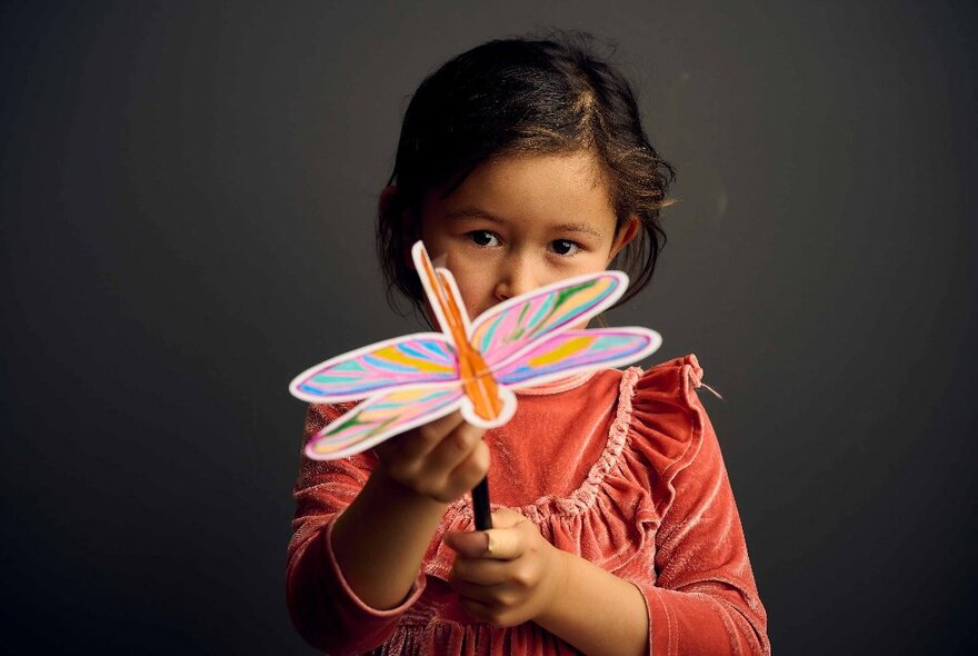 A young child in an frilled orange dress, holding a paper butterfly on a stick.