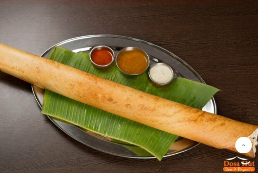 Rolled up dosa on palm leaf with condiments, on silver tray.