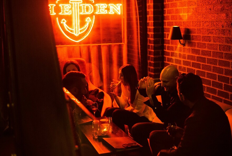 People drinking in a red-lit bar with neon 'The Den' sign, brick wall and lamps.