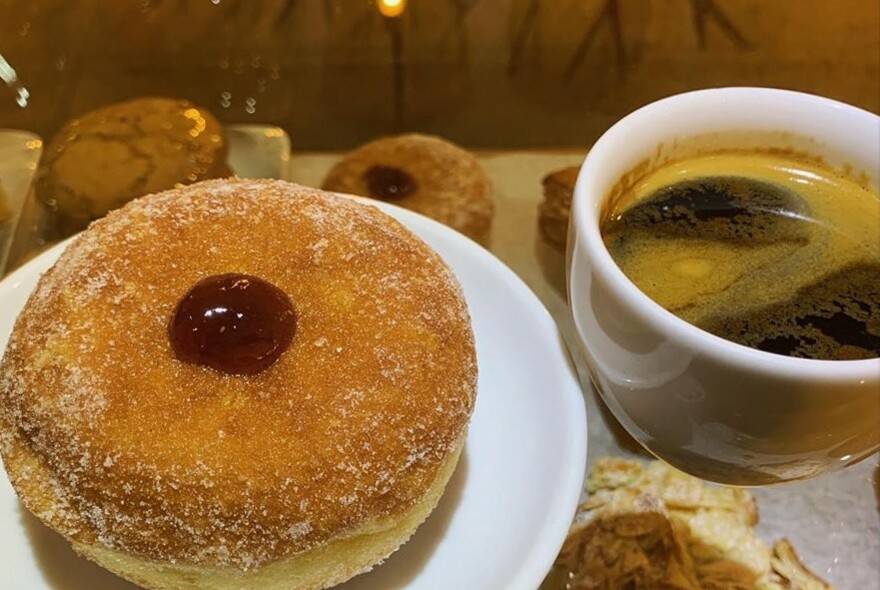 Close-up of a cup of black coffee with a jam doughnut in the foreground.