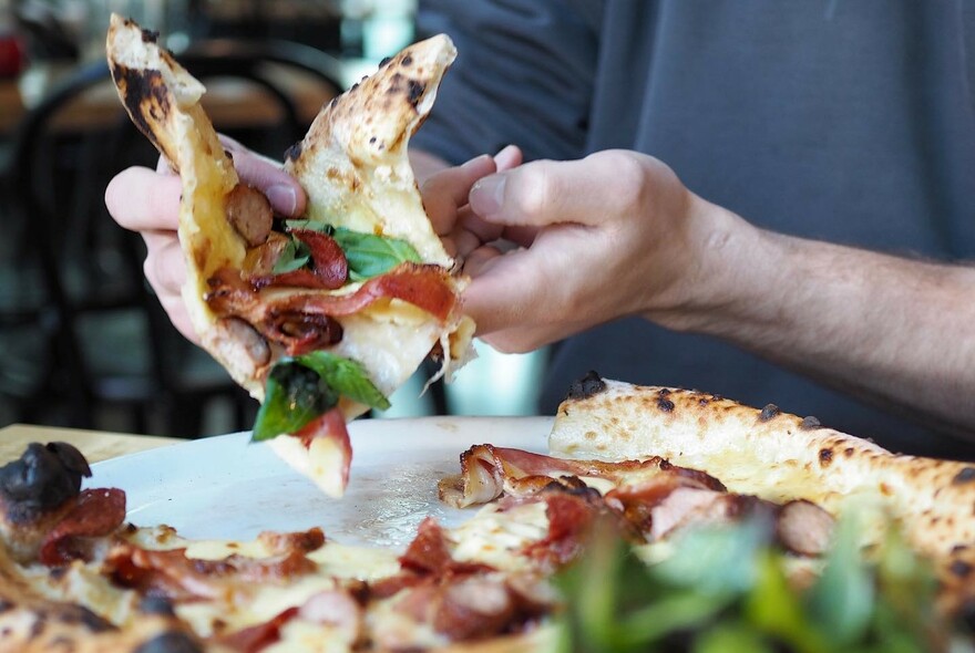 Hands folding a slice of pizza topped with ham.