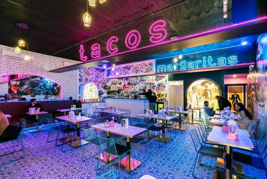 The interior of a Mexican restaurant with empty tables and a large pink neon sign saying 'tacos'.