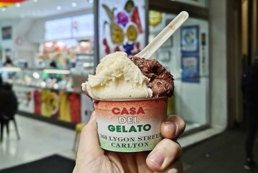 A hand holding up a tub of Casa del Gelato with a spoon in it outside the store. 