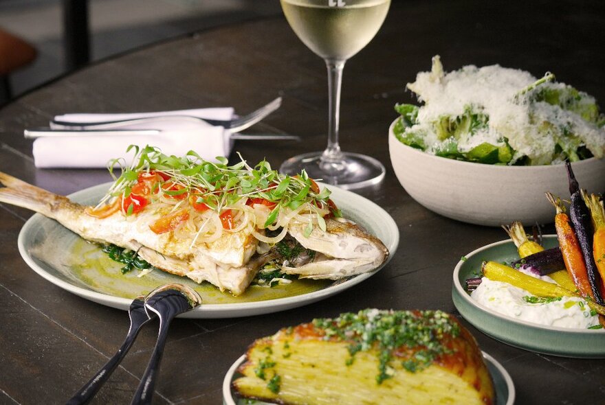 Dining table with plates of food including a whole baked fish, a green salad, and a glass of white wine.