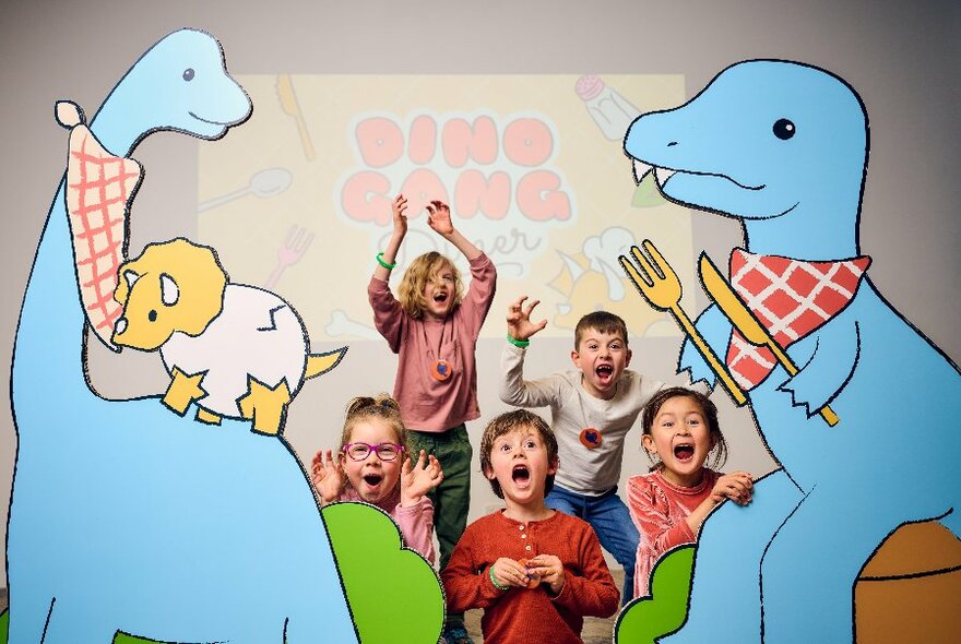 Cartoon dinosaurs with bibs and cutlery with real life kids in the middle, mouths open.