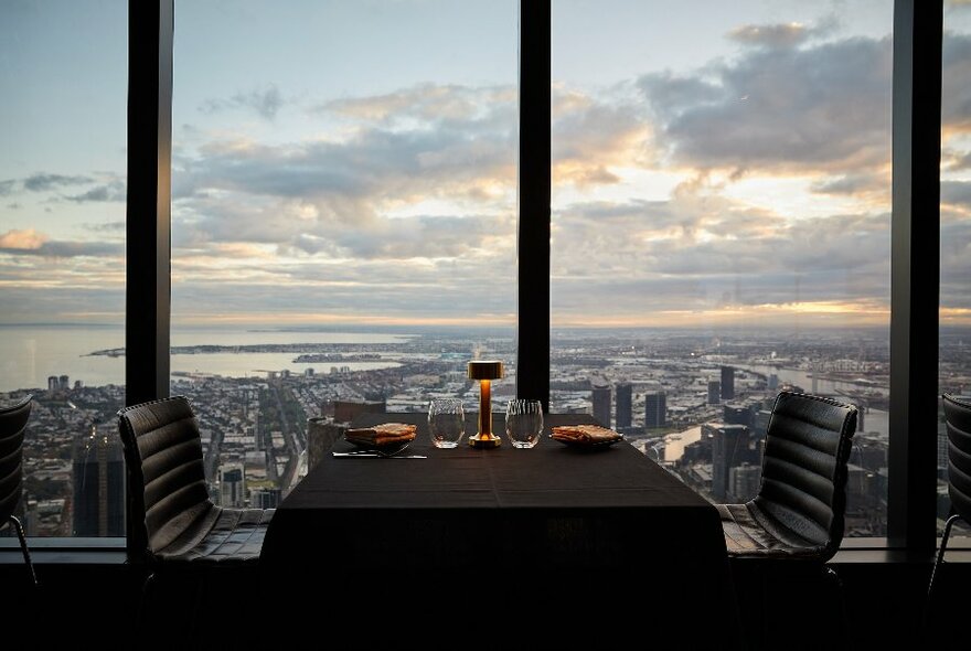 Interior view of Eureka 89 with a dining table and two chairs in the foreground positioned against a large window, with a panorama of Melbourne city and the bay visible in the background.