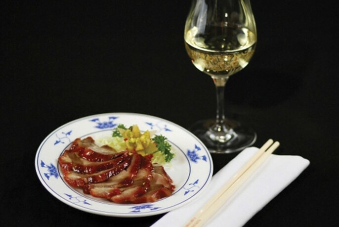 Black table with plate of roast pork, chopsticks and glass of white wine.