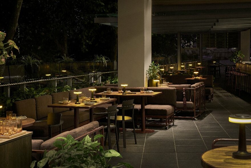 View at night of an outdoor terrace area with tables, chairs, couches and greenery in planter pots.