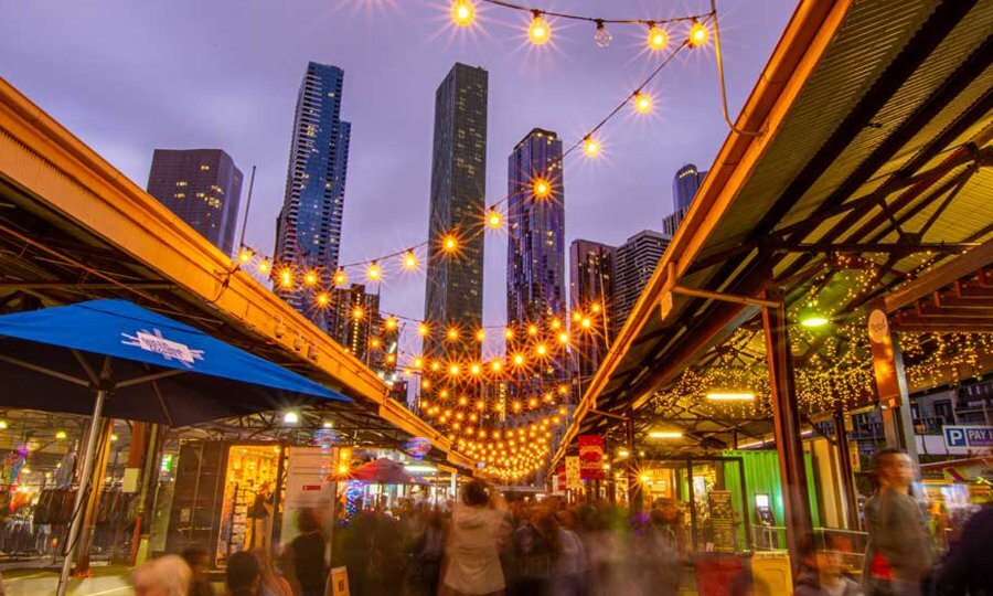 A busy open-air market in the city at night with festoon lighting strung between the sheds.