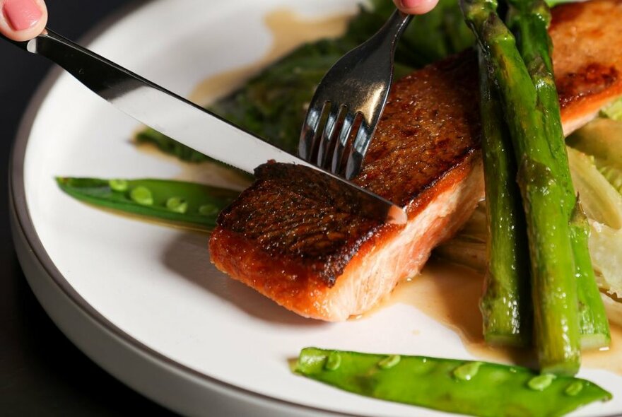 Cutlery cutting into a grilled salmon fillet on a white plate, with some green vegetables to the side.