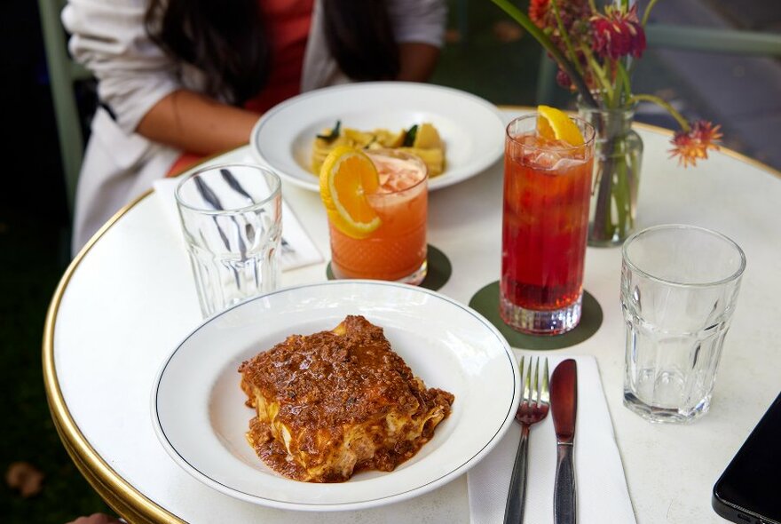 A plate of lasagne on a restaurant table with cocktails.