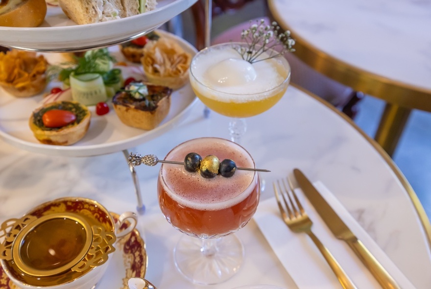 Two different cocktails next to a cake platter on a marble table set with gold cutlery. 