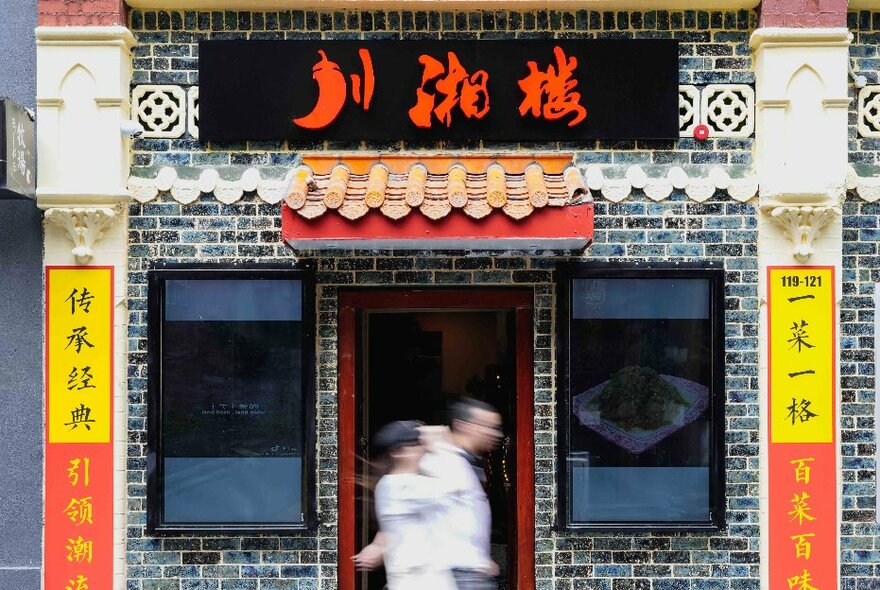 The exterior of a Chinese restaurant with Chinese writing in the top and sides and traditional looking motifs - two people walking in blurred motion out the front. 