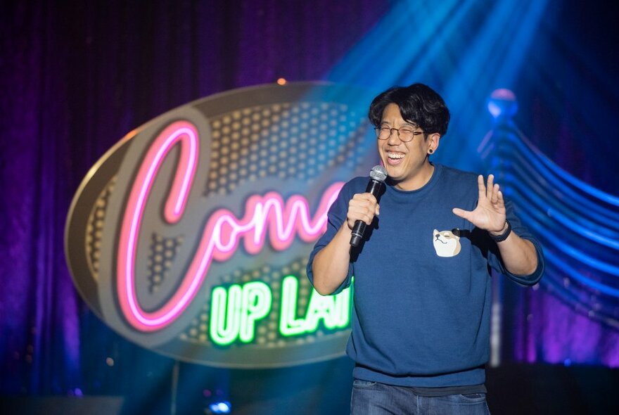 A smiling comedien in a blue t-shirt and jeans holding a microphone on stage in front of a neon 'Comedy Up Late' sign.