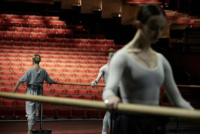 Three ballet dancers on a stage in training poses holding a barre, with red empty theatre seats behind them.