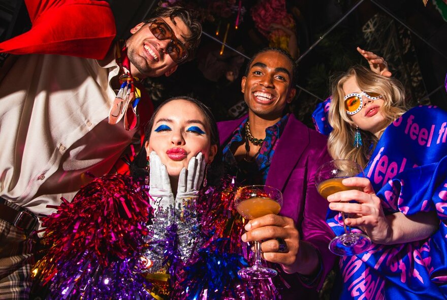 Four people dressed up, smiling, dancing and  having a fun time drinking cocktails, with sparkling decorations around them.
