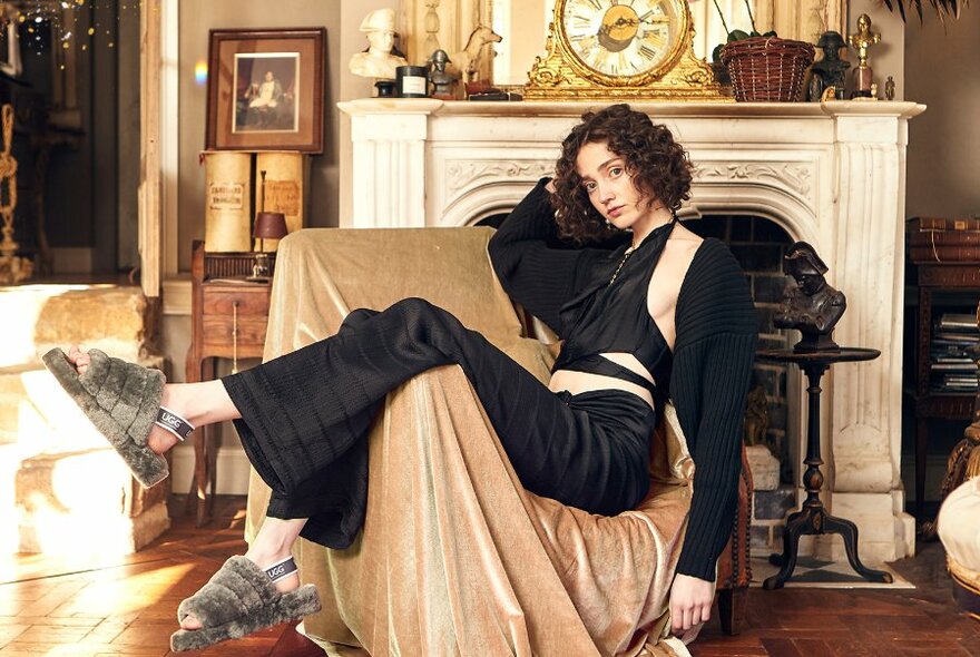 Model reclining slouched in an armchair in an opulent lounge setting, wearing sandals made of sheepskin on her feet.
