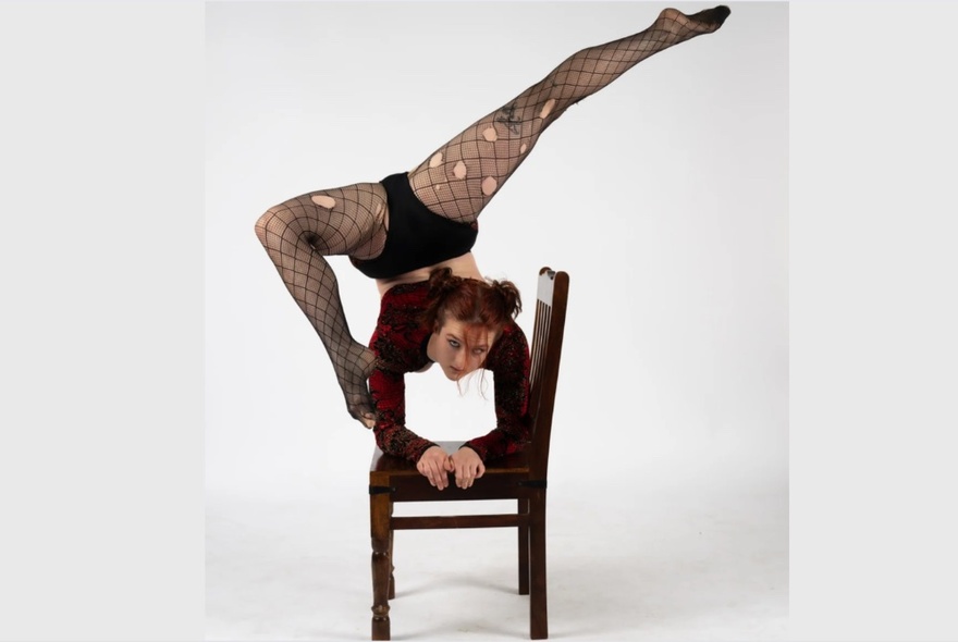 A burlesque artist performing an elbow stand on a chair with legs in the air.