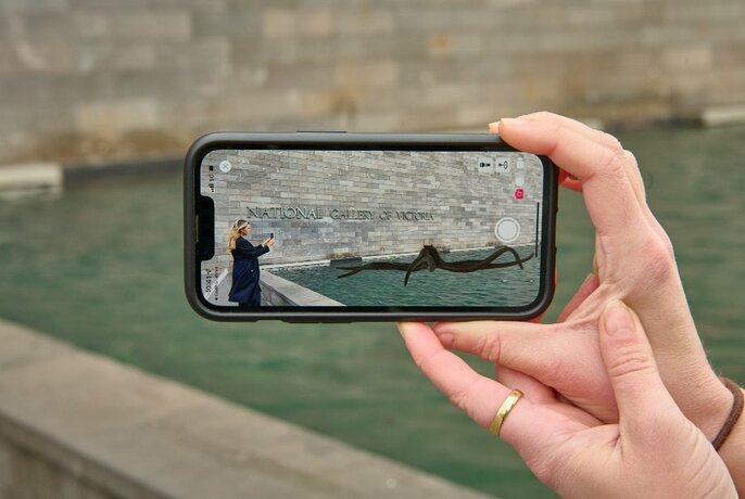 A hand holding a smartphone photographing a person in front of an art installation outside NGV.