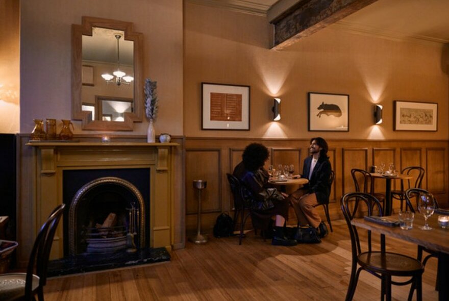 Two people seated in an art-deco style pub next to a fireplace.