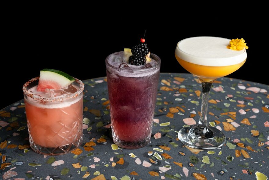 Three coloured cocktails, with garnishes, on a patterned table top.
