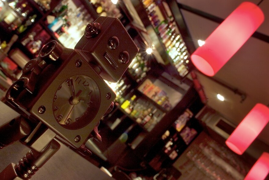 Silver robot clock in front of a backlit bar with bottles and pink vintage lights.