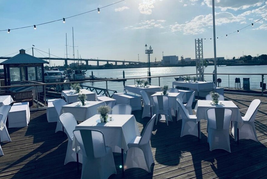 White dining tables and chairs on a wooden deck with the Westgate Bridge and water in the background.