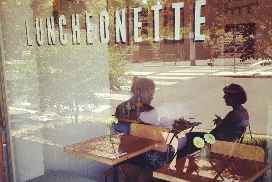 People sitting at an outdoor table reflected in Luncheonette's front window, with signage.