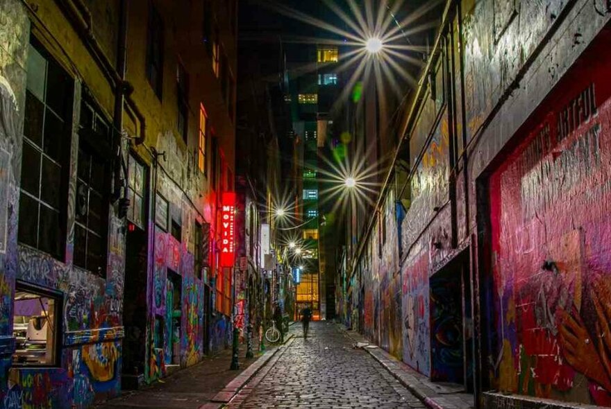 A person walking down graffiti-covered Hosier Lane at night with glowing lights.