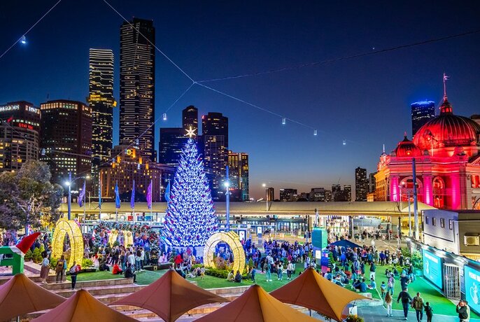 View over the plaza at Fed Square with Christmas decorations including an illuminated tree, crowds, with city skyscrapers and the red-lit Flinders Street Station Facade visible in the background.