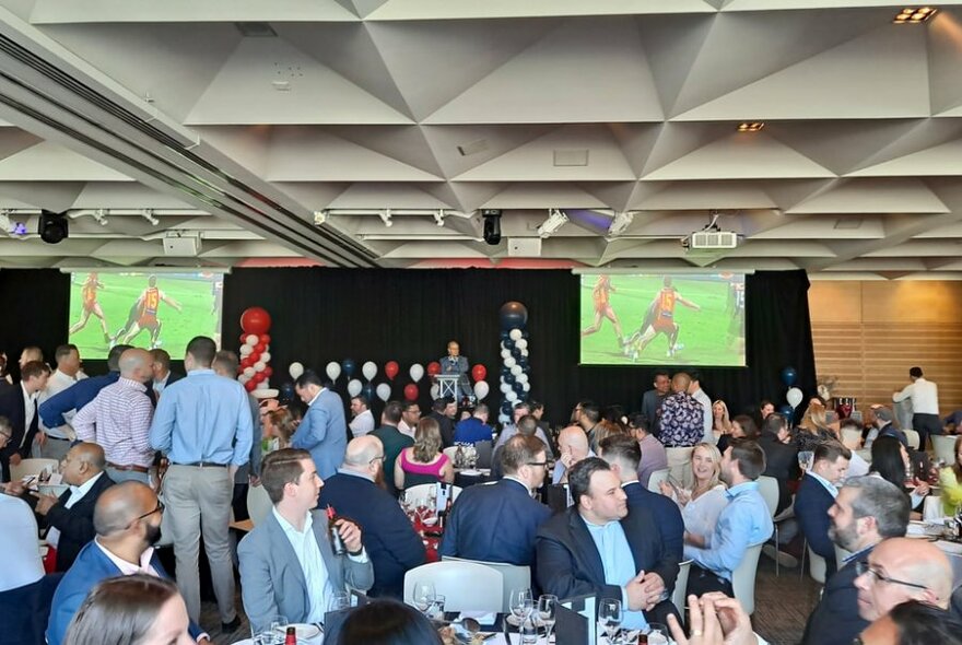People sitting or standing around at round tables mingling and talking inside a large room, with two large television screens showing sport on a read wall.