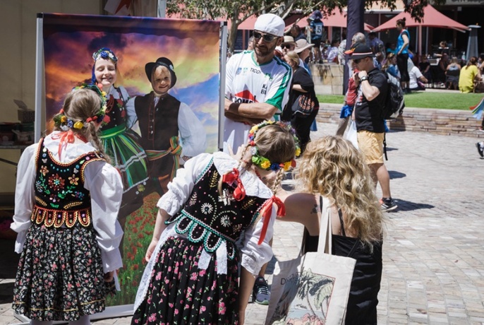Two children in traditional Polish dresses beside adult bopping down, seen from behind, looking at two children with faces sticking out of cut out photo board.