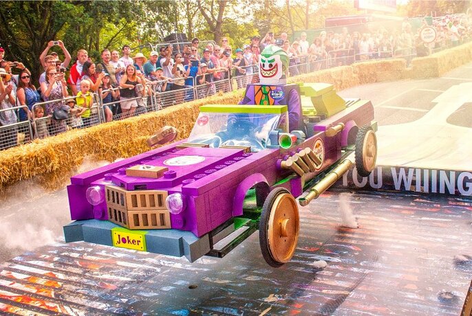 Person steering an elaborate purple billy cart on a street track, past onlookers standing behind a row of hay bales.