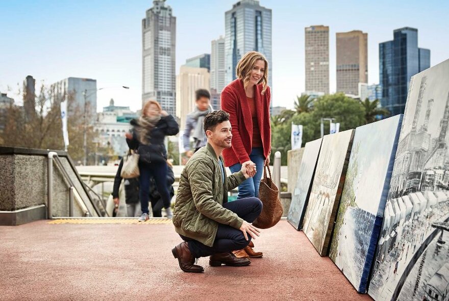 Couple looking at large paintings on display on a footpath, with Melbourne skyline in the background.