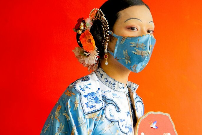 Top half of a woman wearing a traditional Chinese embroidered garment, with a decorative floral piece in her tied back hair, and a patterned cloth Covid-era face mask over her mouth and nose.