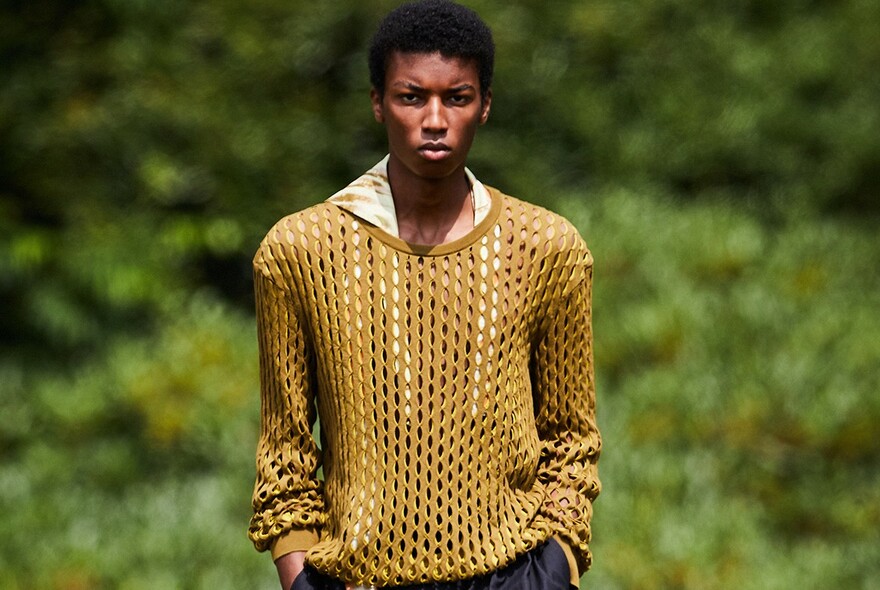 Male model wearing loose yellow knitted sweater, with blurred green foliage in background.