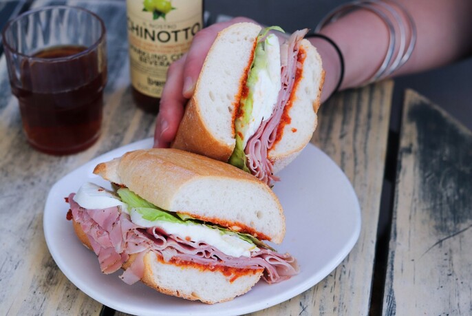 A woman holding a panini filled with sliced meat, cheese, lettuce and condiments. A glass and bottle of soft drink is also on the table. 