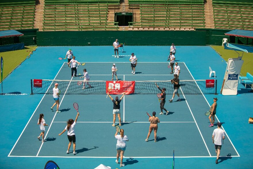 Many people on a blue outdoor tennis court holding racquets.