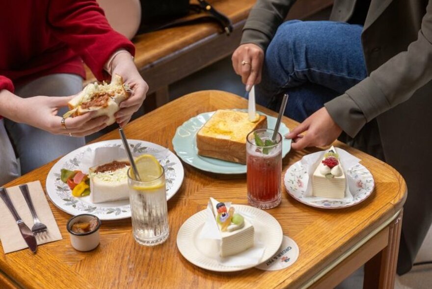 Wooden table with two slices of cake on separate plates, two glasses of soda, girl buttering thick toast, another girl is holding a sandwich.