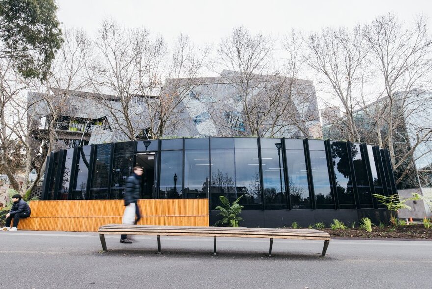 Fed Square Greeline Project Hub building surrounded by bare winter trees and empty public bench, one person walking past.