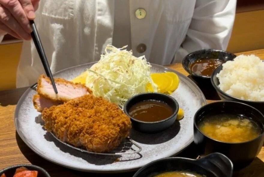 Person in white shirt picking up piece of fried katsu chicken with chopsticks. Plate of katsu chicken is surrounded by sauces.