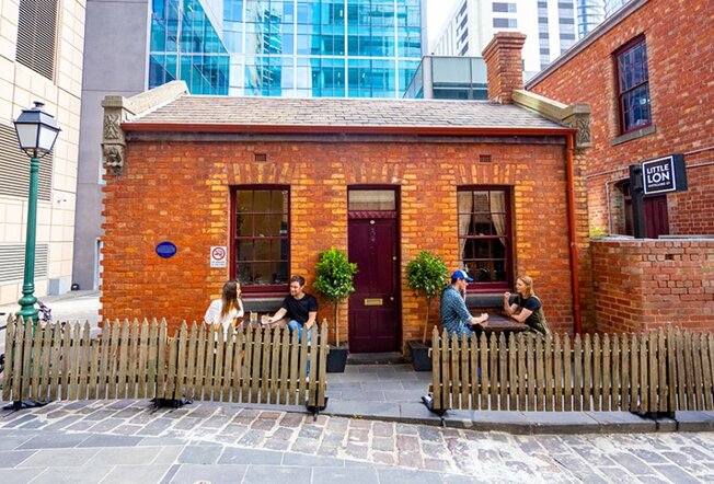 A small brick building in the city with people drinking outside behind a picket fence.
