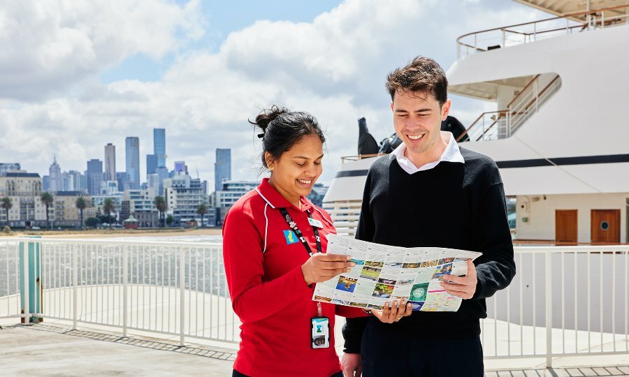 A tourism employee standing beside a cruise shop and sharing a map with a tourist. 