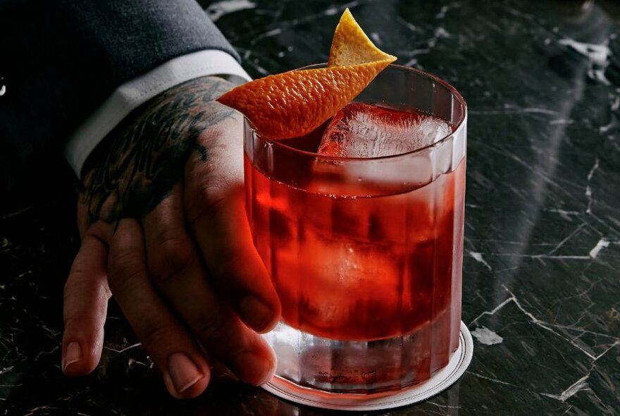 Hand offering red cocktail in highball glass across black and white marble-look surface.