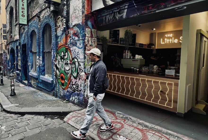Exterior of a walk-up small cafe kiosk in Hosier Lane, with a person walking past, street art on the walls of the buildings in the lane.