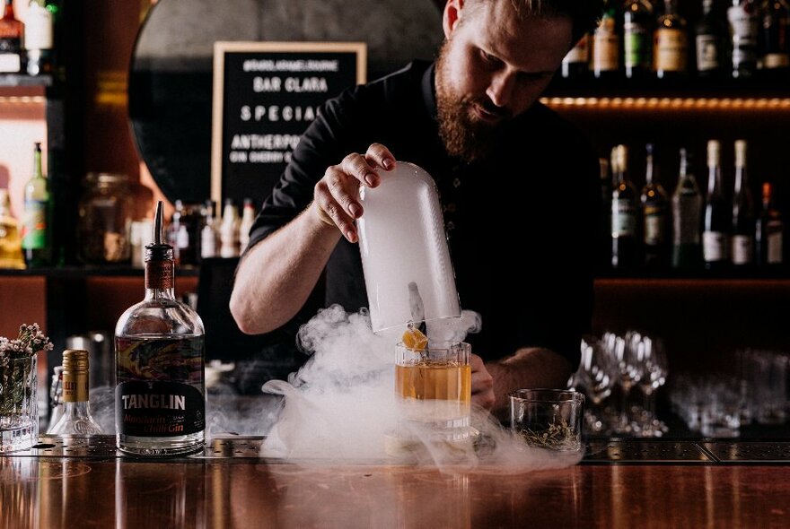 Cocktail being created and presented at a bar, with a person behind the bar and shelves of bottles visible.