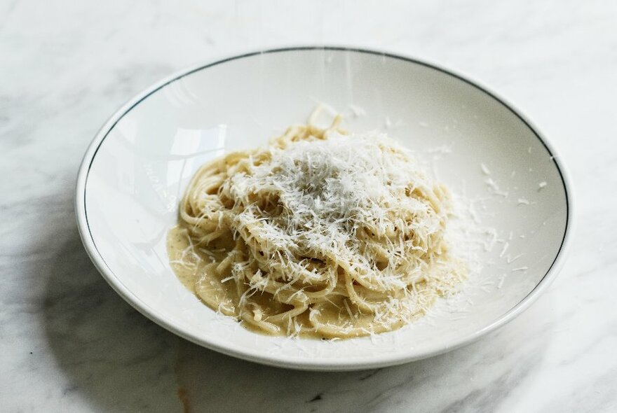 A white bowl of cacio e pepe with cheese on top on a marble table.