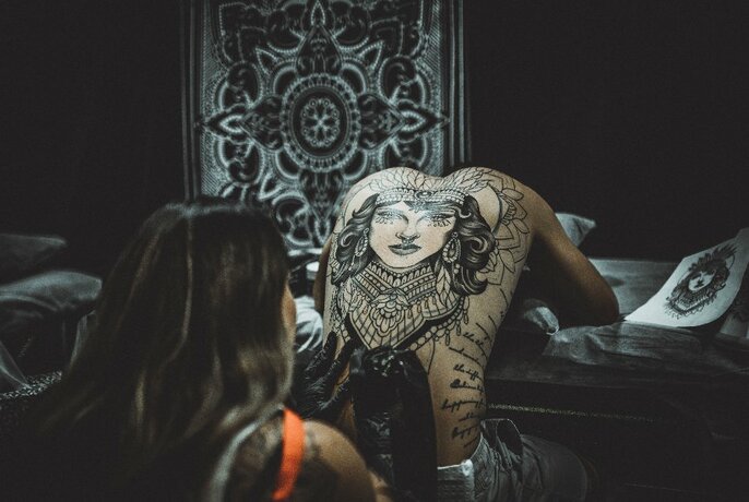 A tattoo artist tattooing the back of another person in a darkened room. 