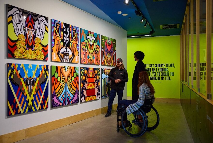 Two people, one in a wheelchair and one standing, looking at a wall of colourful art while a third person explains it to them.